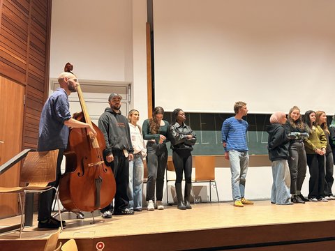 Das Bild zeigt einige Personen auf der Bühne des Audimax die etwas präsentieren. Einer hält ein großes Streichinstrument.