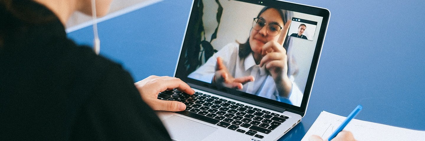 Eine Frau sitzt an einem Tisch mit Laptob und Stift und Zettel, auf dem Laptop ist eine andere Frau im Videochat zu sehen und sie reden miteinander