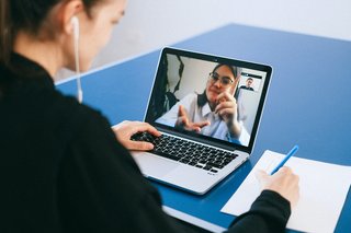 Eine Frau sitzt an einem Tisch mit Laptob und Stift und Zettel, auf dem Laptop ist eine andere Frau im Videochat zu sehen und sie reden miteinander