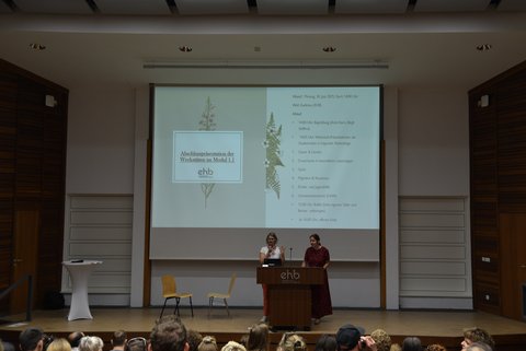 Zwei Personen stehen auf der Bühne und im Hintergrund ist ein Plakat zu sehen.