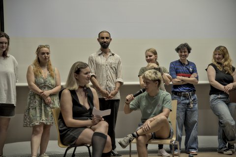 Zwei Menschen sitzen im Vordergrund auf Stühlen und andere Menschen stehen im Hintergrund und hören ihnen zu.