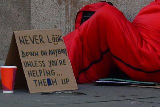 Obdachloser in Schlafsack mit Schild "Never look down on anyone, unless you're helping them"