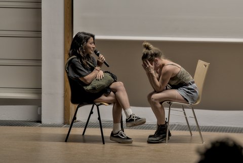 Zwei Menschen sitzen auf Stühlen. Die rechte Personen spricht ins Mikrofon und die rechte sieht bestürzt aus.