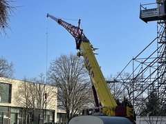 Gelber Lastkran mit Transportseil, im Hintergrund links der Querriegel E-Gebäudes der EHB