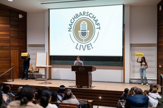 drei Studierende stehen auf einer bühne vor einem projektor, auf dem „nachsbarschaft live“ steht. einer von ihnen präsentiert hinter einem podium, während die beiden anderen schilder mit der Aufschrift „lachen“ hochhalten