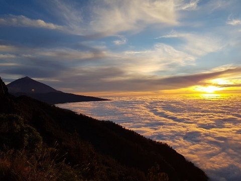 Blick von einem Berg. Der Ausblick ist so weit oben, dass man unten Wolken sieht. Die Sonne geht unter