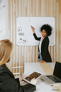 Eine Frau zeigt auf ein Whiteboard während sie etwas erklärt.