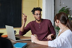 Ein Mann und eine Frau sitzen einander zugewandt an einem Tisch vor sich einen Laptop