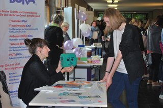 Foto mit Menschen an einem Ausstellertisch vor Informationsplakaten