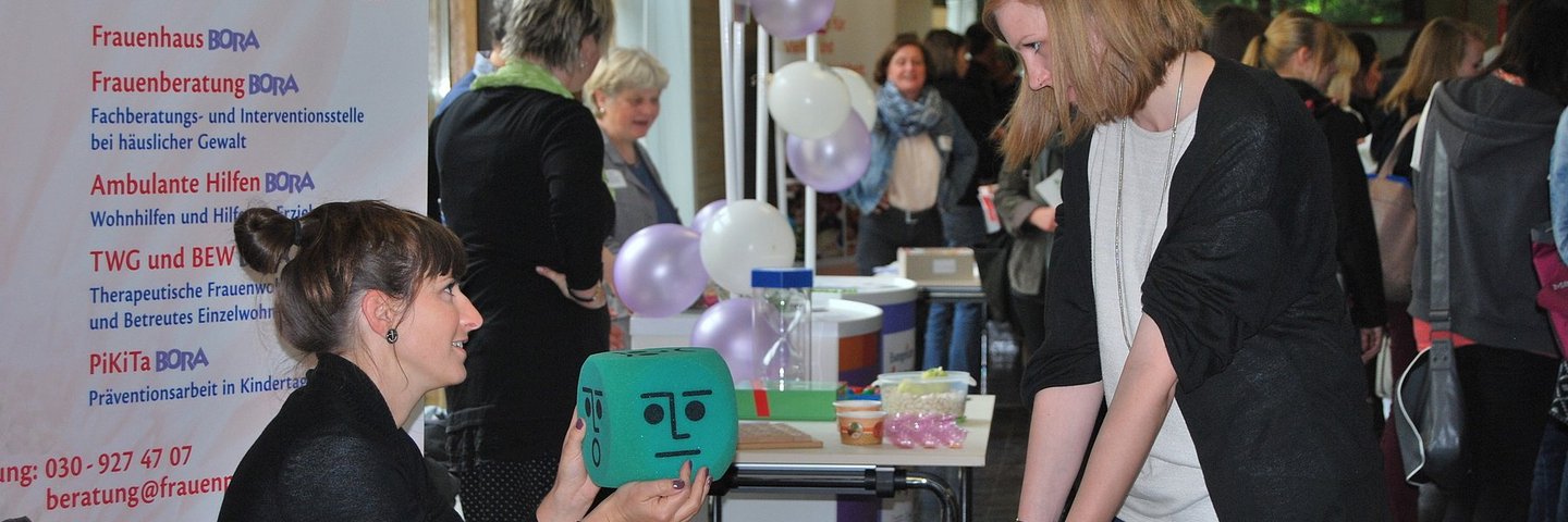 Foto mit Menschen an einem Ausstellertisch vor Informationsplakaten