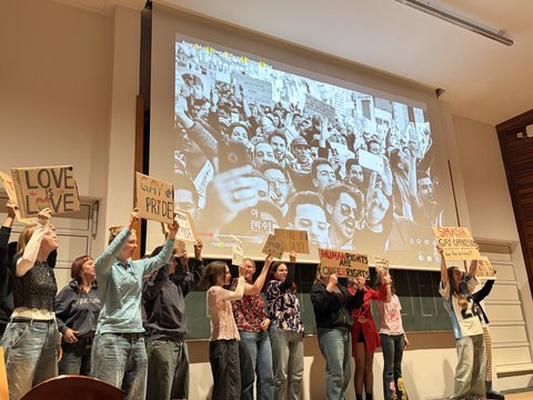 Das Bild zeigt einige Personen auf der Bühne des Audimax die etwas präsentieren und Plakate hochhalten.