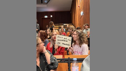 Das Bild zeigt Studierende die ein Schild hochhalten auf dem steht "Hört auf, uns unsichtbar zu machen".
