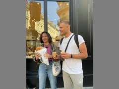 Zwei Menschen, eine männlich gelesene und eine weiblich gelesene stehen vor einem Schaufenster und haben Kaffe und Gebäck in der Hand